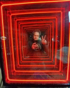 Vibrant red neon square tunnel art illuminated with a man making a peace sign in mirror reflection.