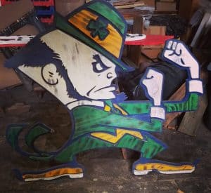 Vivid painted wooden cartoon-style leprechaun figure with flexed arm, green suit, and hat, handcrafted by Chris Knight Creations.